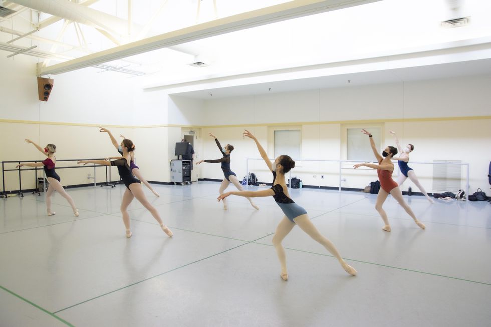 In a spacious, light-filled dance studio, two lines of teenage female ballet students do a tendu derriere effacu00e9 in pliu00e9 on their right leg with their arms in third arabesque. They each wear a leotard in various colors, pink tights and pointe shoes.