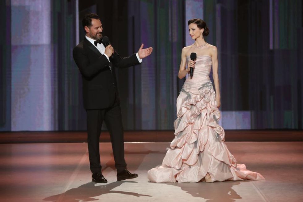 A man in a tux stands on a stage gesturing at Zakharova, in a long pink gown, microphone in hand.
