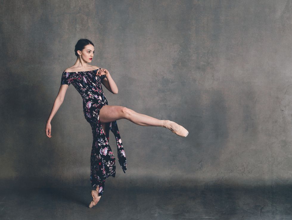 Dolan wears a long floral dress, and stands with on point with her left leg in front attitude