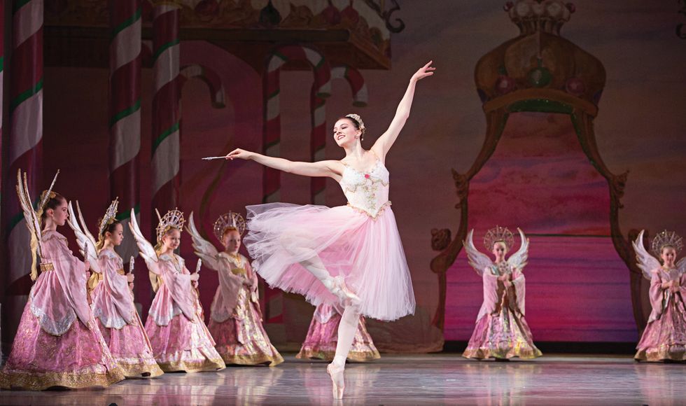 Dolan in a passu00e9 on pointe holding a star wand, in a long pink tutu, against a backdrop of children angels dressed in pink.