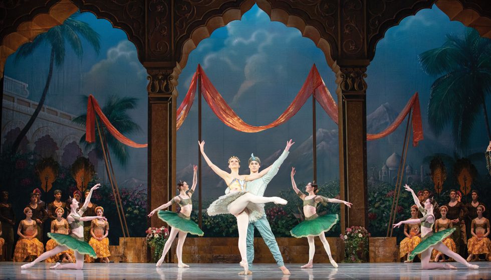 Dolan in center stage supported by Baca, she stands on point in a front attitude with her arms in the air, in a pale green and yellow tutu