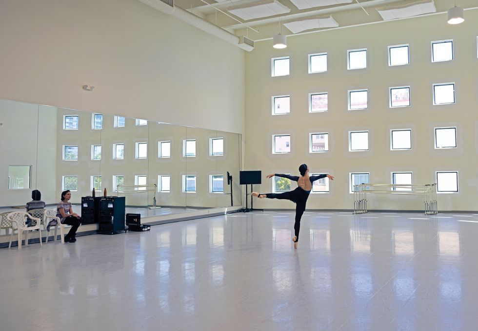 Morgan is seen from behind in an arabesque on pointe. Lopez sits in a white plastic chair in front of the mirror, watching her.