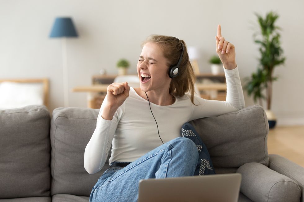 A young woman sits on her coach with headphones in and a laptop in front of her, singing expressively.