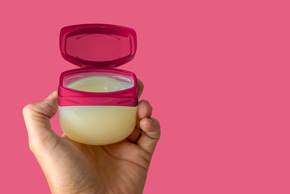 The left hand of a young female person holds a container with skincare cream over colored clean background.