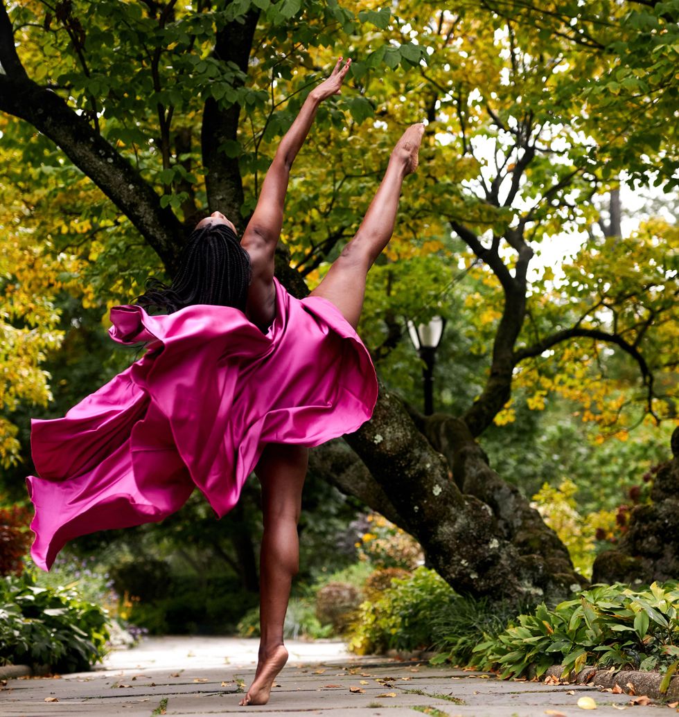 A Black female dancer in a wooded park and draped in fuchsia satin faces away from the camera and does a devu00e9loppu00e9 to the side with her right leg.