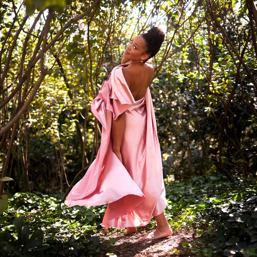 A woman draped in pink satin stands in a wooded glen with her back towards the camera and turns her head over her left shoulder.