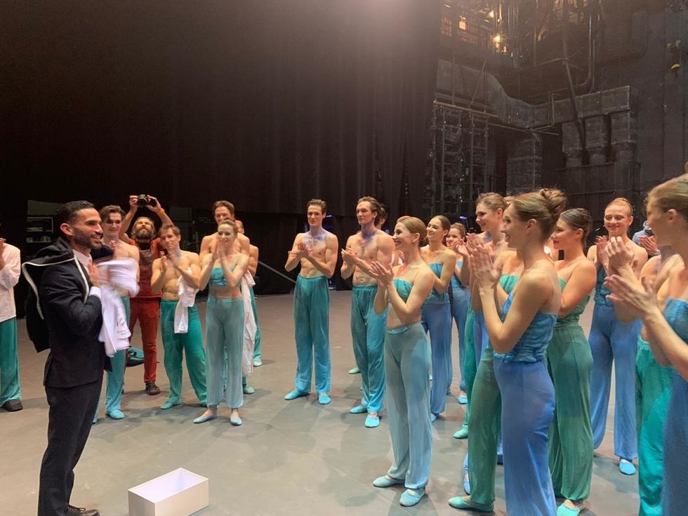 A large group of dancers wearing blue costumes stand backstage around a choreographer in a suit, clapping towards him in congratulations.