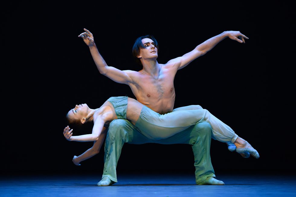 A bare-chested male dancer squats and balances the body of a ballerina costumed in blue, who lays across his legs.