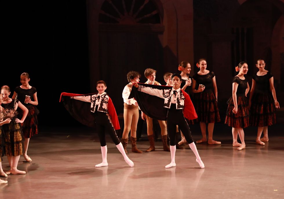 Students in performance. Two boys in torero costumes are center stage, holding one side of the red cap out to the side with the opposite leg in tendu. Crowds of other dancers in Spanish themed costumes are behind them.