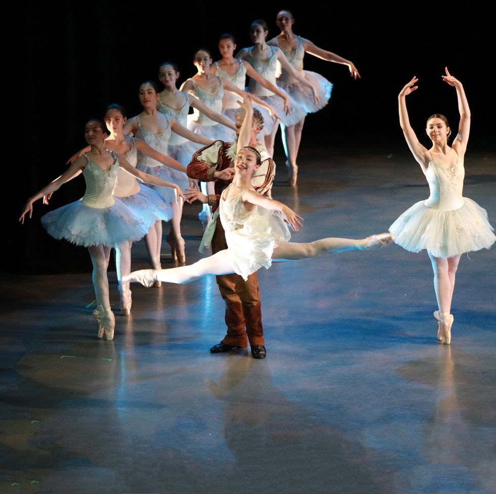 Students in performance. Rows of girls in white and blue romantic tutus, en pointe in fifth position with their arms to the sides. A girl center stage is mid-leap, in a flowy dress.