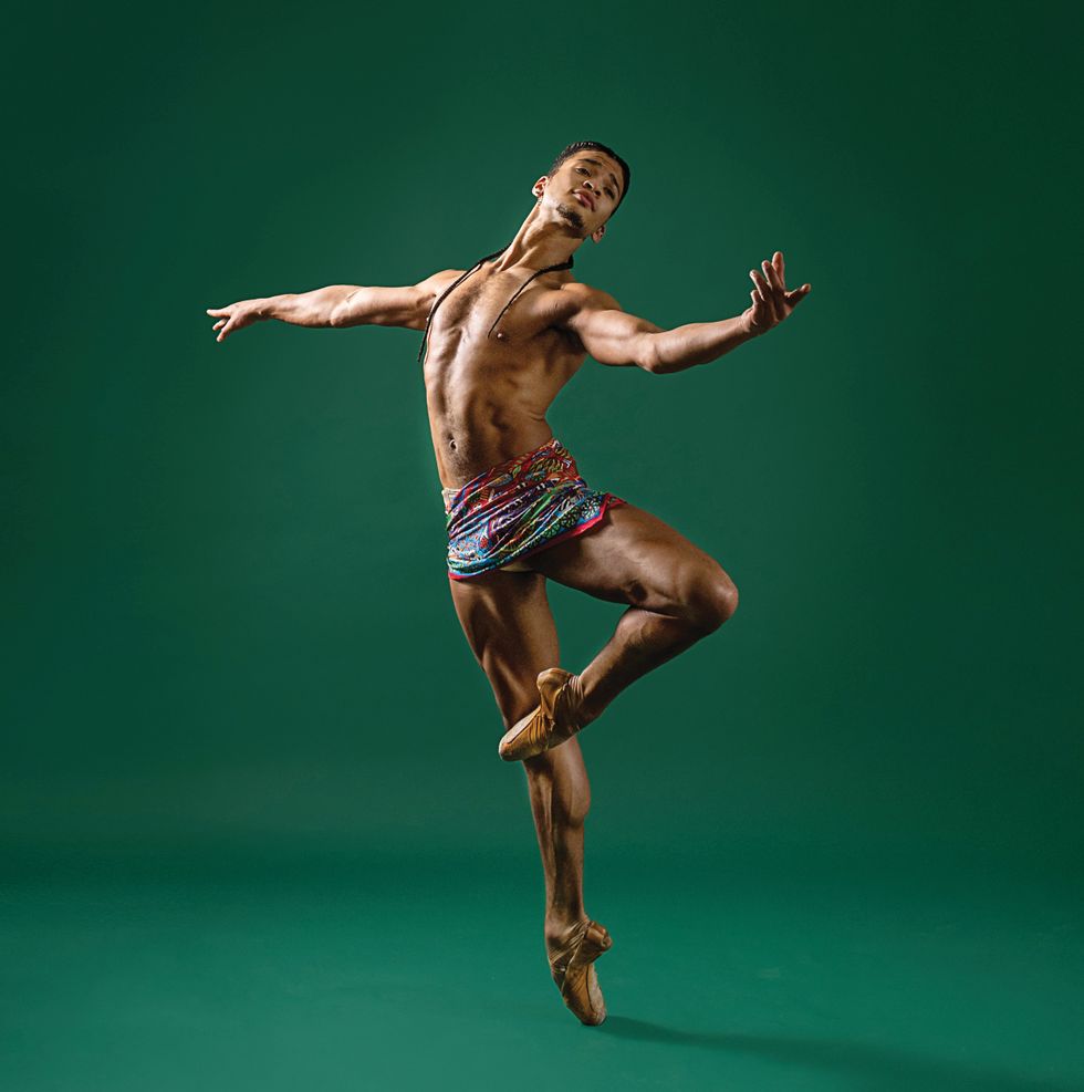 Hayes in a short colored wrap skirt and pointe shoes in a passu00e9 with his arm extended to the side