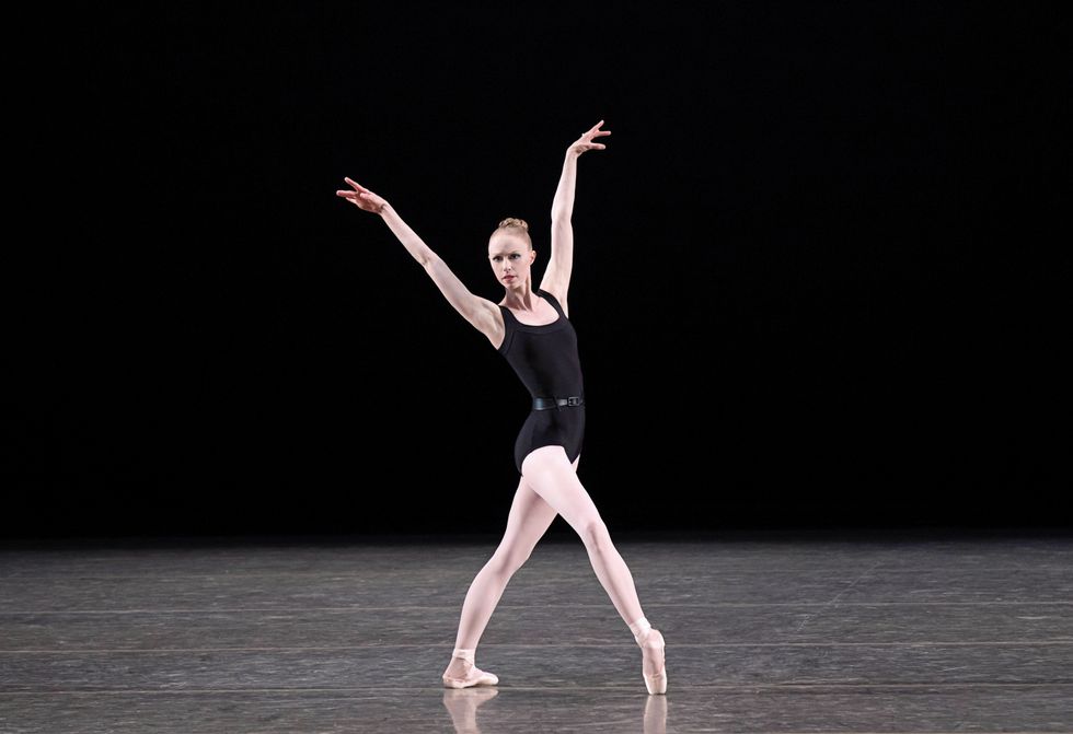 Reichlen, in pink tights and a black leotard with a black belt, stands in a wide fourth position with her arms in the air.