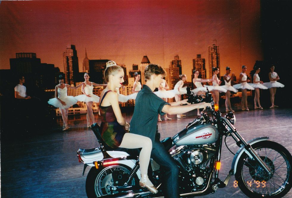 Schull and Stiefel sit on Stiefel's motorcycle behind the scenes. Schull wears a dark red dress, pink tights, pointe shoes, and her hair in bun, with a bedazzled bun cage. Stiefel wears black jeans and a black button down. In the background, ballerinas in white tutus pose in a line.