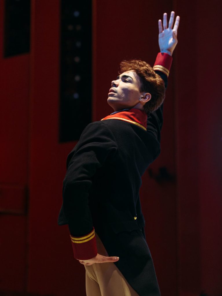 Close up of Victor Caixeta in rehearsal in a regal jacket with one hand on his hip and the other flung above his head.