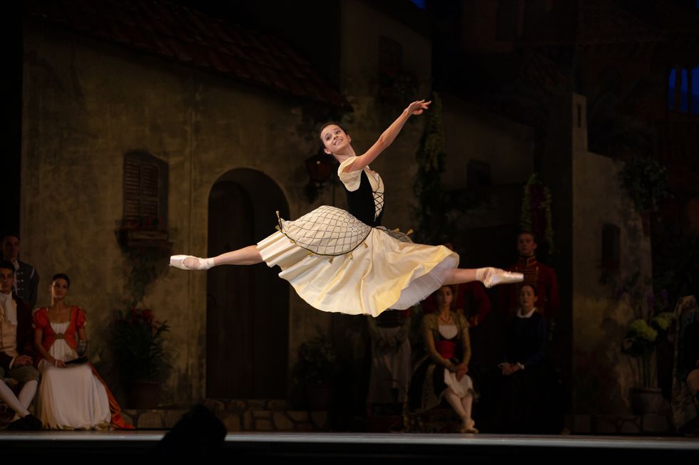 Maria Khoreva, in a yellow skirt and black bodice, leaps across the stage, smiling at the audience.