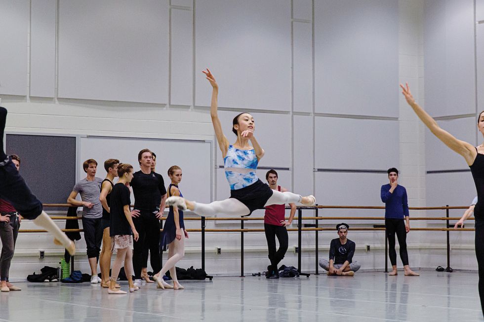 Nagahisa in a grand jeté in the middle of a rehearsal studio, wearing a blue and white leotard, black shorts and pink tights.