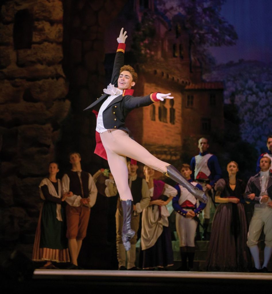 Caixeta, in brown tights and boots a black jacket jumps in the air in rehearsal onstage, smiling at the audience.