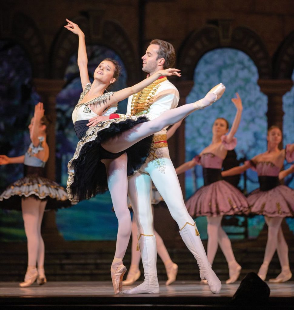 Khoreva, in a black and gold tutu, stands in arabesque on pointe onstage, held around the waist by Zverev, dressed all in white and gold.