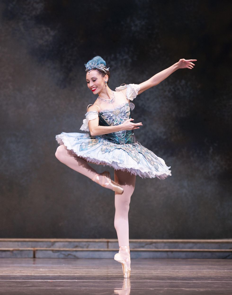 Sarah-Gabrielle Ryan does a piquu00e9 retiru00e9 on her left foot and holds her arms in fourth position en evant, leaning her upper body over to look towards her right leg. She wears an ornate blue tutu with off-the-shoulder sleeves, a blue feathered headress and pink tights and pointe shoes.