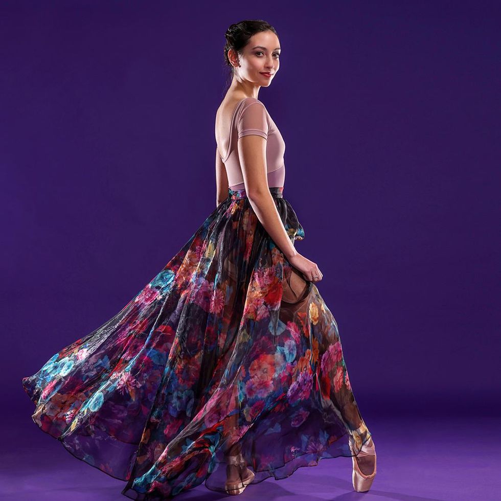 Standing in front of a bright purple backdrop, Sarah-Gabrielle Ryan wears a pink cap-sleeved leotard and long chiffon skirt with a bright floral pattern. She faces profile to her left, props her right foot up onto her pointe shoe and looks back toward the camera with a close-lipped, confident smile.
