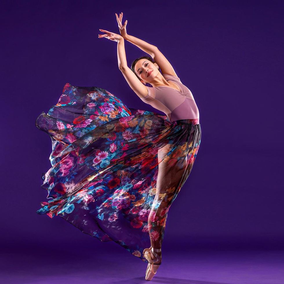 Sarah-Gabrielle Ryan poses in sixth position on pointe. Her legs face profile while her upper body twists front and leans back and her arms raise high above her head with wrists together. Standing in front of a purple backdrop, she wears a pink leotard and long, brightly colored floral chiffon skirt that billows dramatically high behind her in the wind