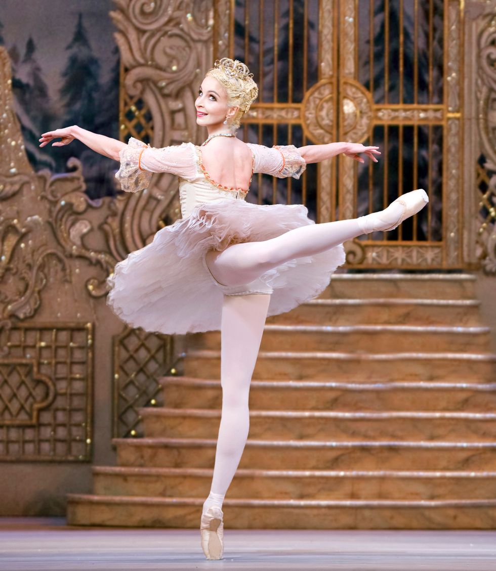 Laurent Cutherbertson, wearing a light pink and gold tutu, blond wig and gold tiara, balances in attitude facing the upstage right corner, looking back towards the audience with a smile.