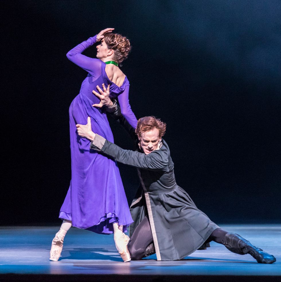 Wearing a long-sleeved purple dress, Lauren Cutherbertson stands in fourth position on pointe, her left hand clutching her forehead. Edward Watson, in a long dark jacket, tights and boots, kneels on the floor and aggressively grabs onto her waist with both hands.