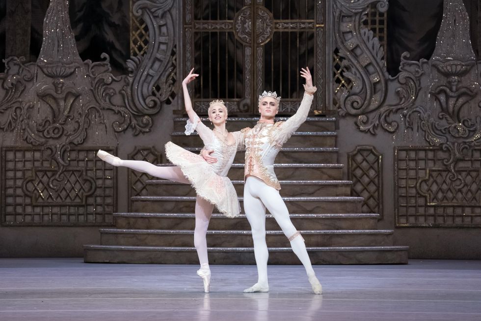 A ballerina in a tiara and light pink tutu, tights and pointe shoes stands in attitude with her right arm up and her left arm around her partner's right shoulder. He stands in tendu and wears a matching jacket and tights and crown.