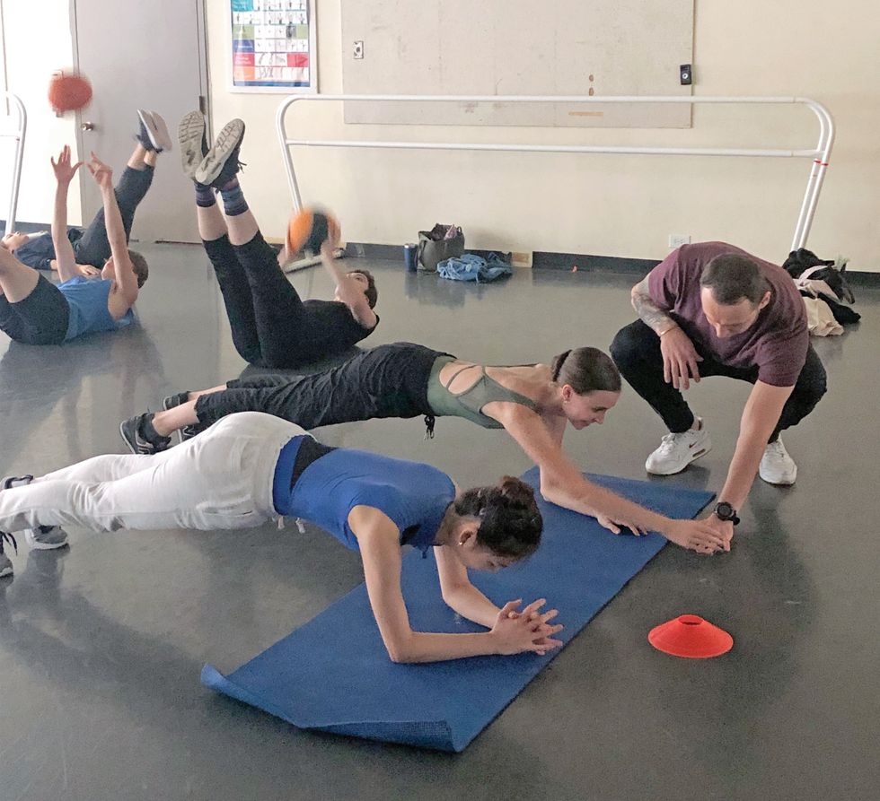 Two women in leotards, sneakers and warm-up pants plank with their forearms on a yoga mat.
