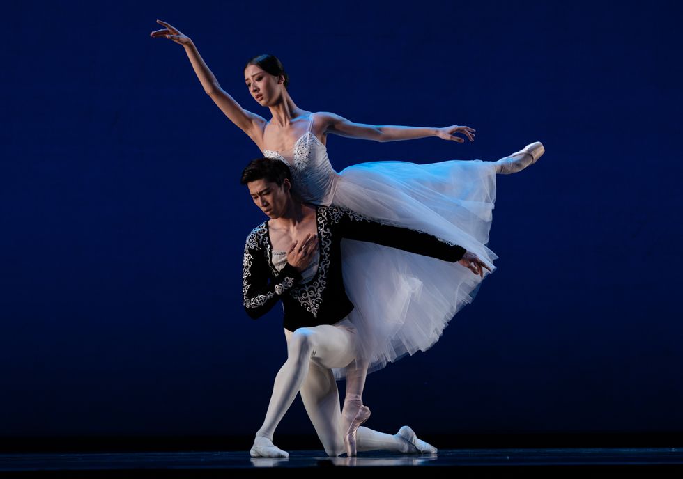 Sicong Wu, in a black tunic and white tights, kneels on his right leg and holds his right hand to his heart. Yunting Qiu, in a long white tutu, balances next to him in first arabesque and leans her torso on his upper back.