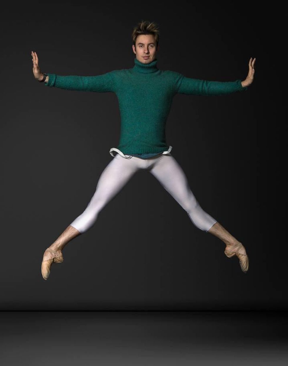 A white male dancer in a green turtleneck sweater, calf-length white tights and tan ballet slippers jumps in second position with his arms out to the side and hands flexed.