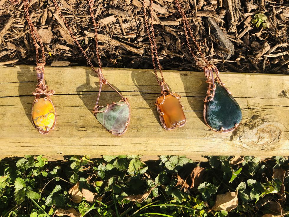 Four copper-wire necklaces with a yellow, light green, orange and dark green stone, respectively, are lined up right to left on a plank of wood.