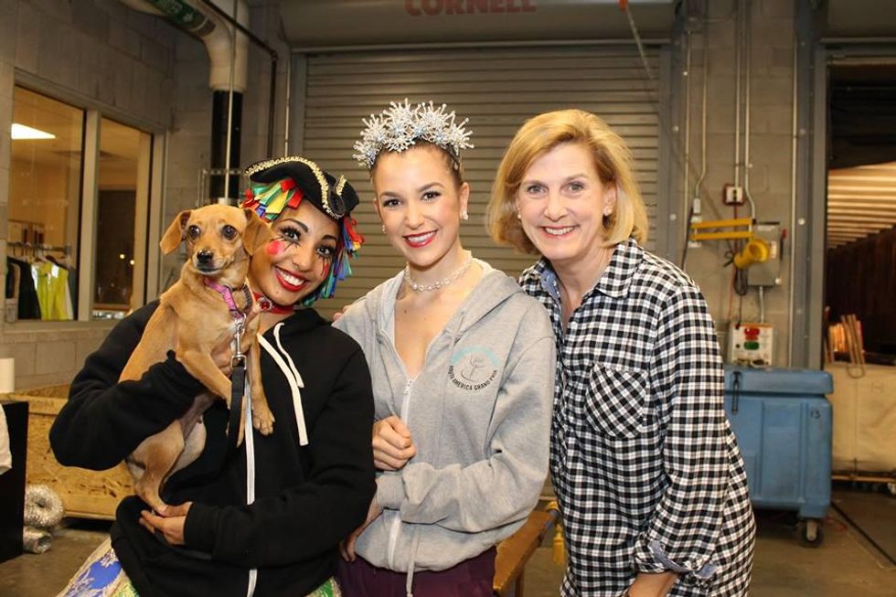 A young Black woman in a black Napoleon hat, black sweatshirt, and stage makeup holds a small brown dog and stands next to a white female dancer in a gray sweatshirt and a sparkly headpiece and an older white woman with a blonde bob and wearing a black gingham blouse.