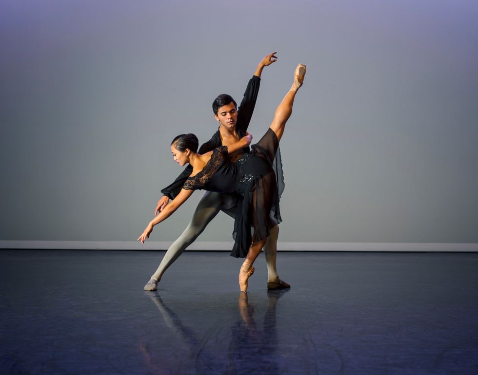 A female college-age dancer in a black sparkly dance dress does arabesque penchu00e9 on pointe while holding onto her partner's torso, who lunges behind her.