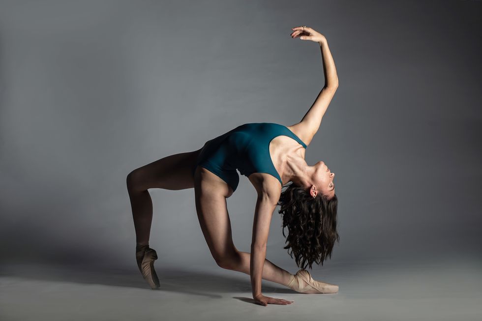 Stefanee Montesantos, in a green leotard and point shoes, pushes over her shoes into a deep backbend, with her left hand on the floor for balance and her right arm reaching up towards the ceiling.