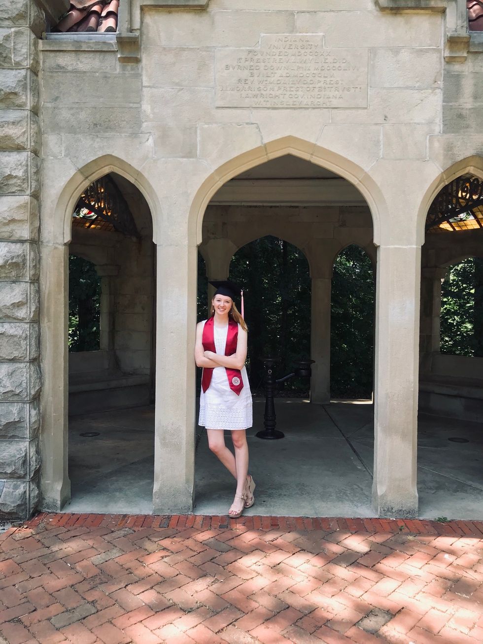 Claudia Rhett, who wears a white dress, graduation cap and sash, leans against a pillar outside on her college campus.