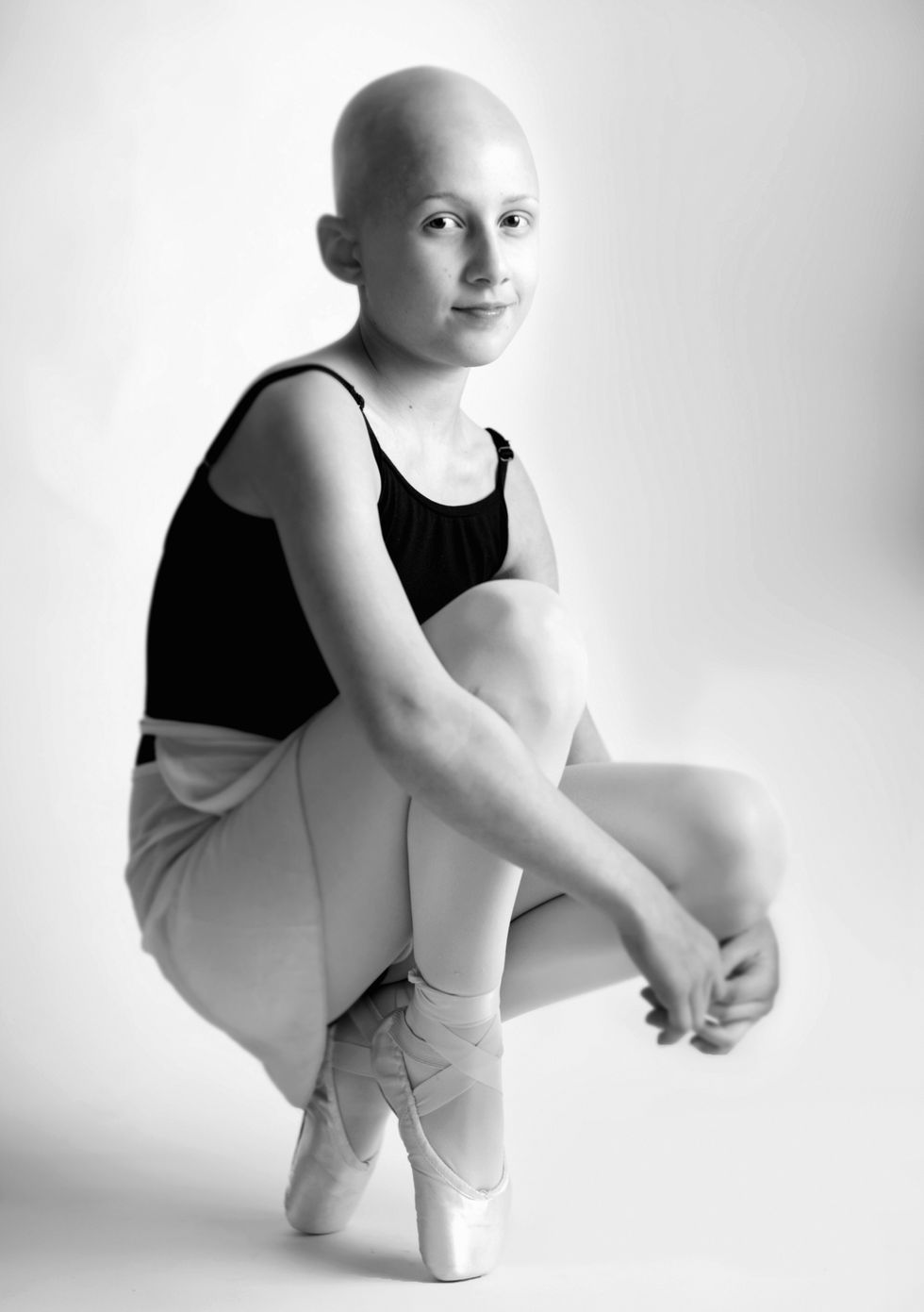 A black and white photo of Stella DiPasquale in a leotard, skirt and pointe shoes, crouched down. Stella has no hair.