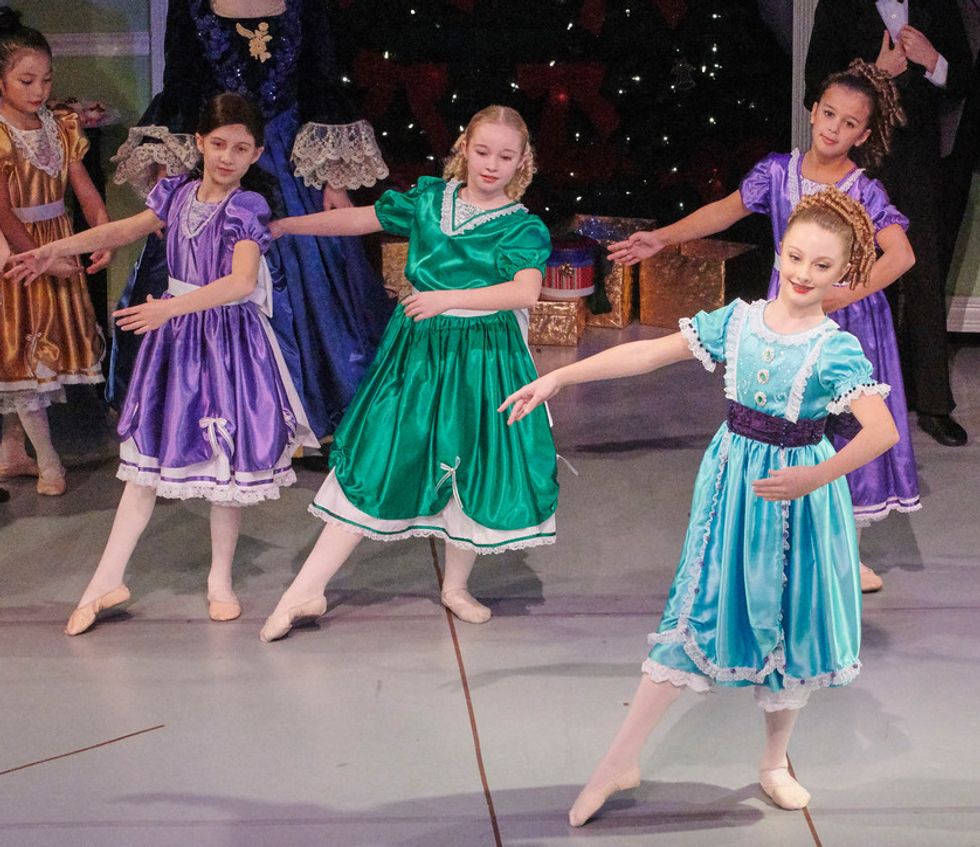 A stage photo of Nutcracker of four young girls dressed in colorful party dresses and soft shoes. Stella DiPasquale wears a purple dress and a brown wig.