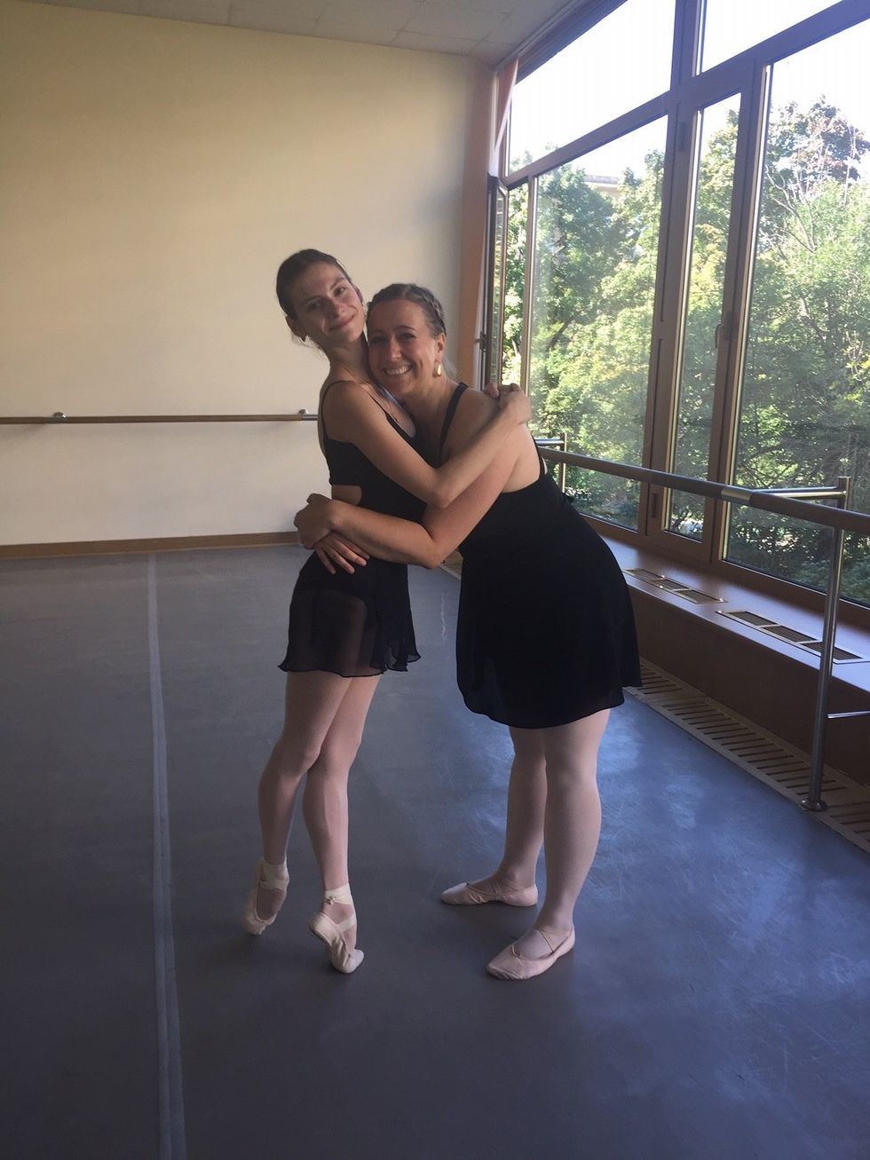 Strakhova (left) hugs Collier while Strakhova stands in relevu00e9. Both women are wearing ballet slippers, pink tights, and black ballet skirts and leotards, while standing in a ballet studio.