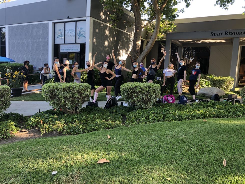 A group of young teenage dance students wearing dance practice clothes and face masks make fun, Broadway style poses in front of a dance studio building, surrounded by lush green grass and landscaping.