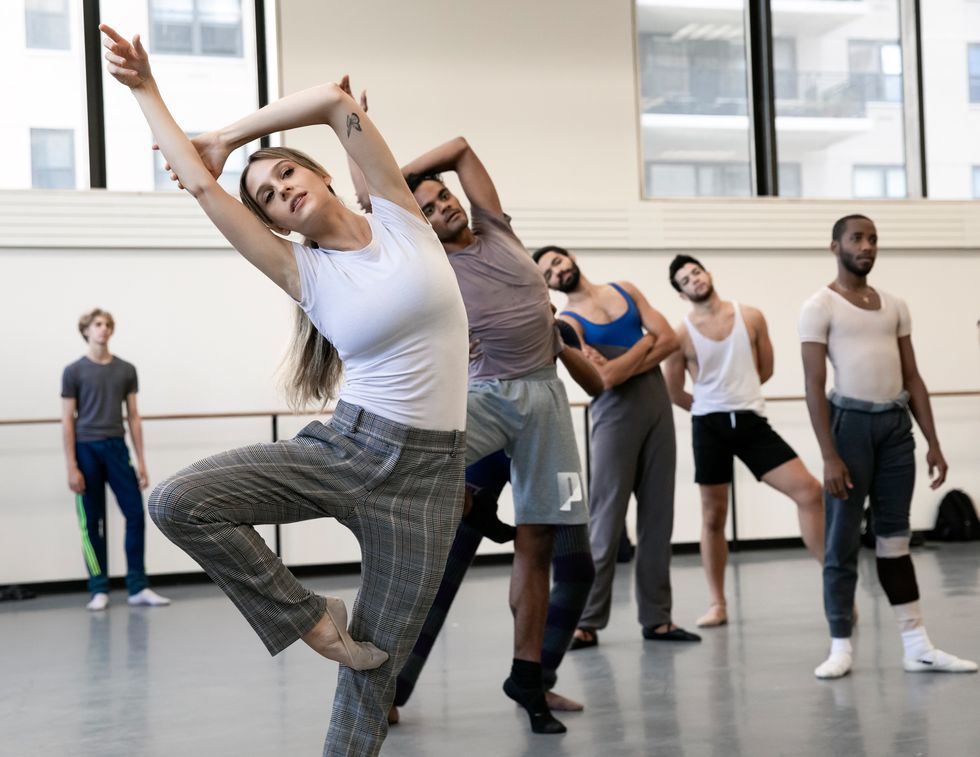 Lauren Lovette, wearing a white T-shirt, gray plaid pants and ballet slippers, does a passu00e9 with her right leg and bends sideways to the right. She lifts her right arm up and grabs her right elbow with her left hand, framing her head with her arms.