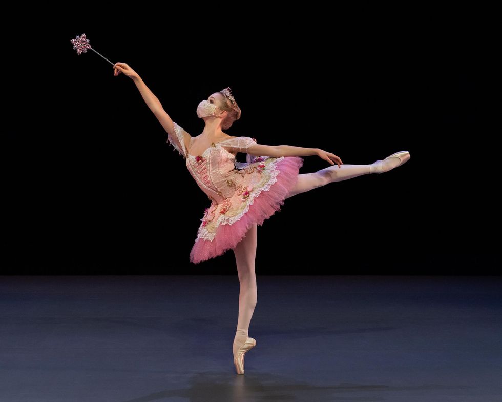 A teenage ballerina holds a sparkly pink wand and does a piquu00e9 first arabesque on her right leg. Costued as the Sugarplum Fairy, she wears a pink tutu with floral and lace details on the skirt and bodice, a sparkly tiara, and pink tights and pointe shoes.