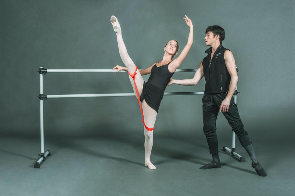 A young white baller student in black leotard and skirt and pink tights and slippers performs du00e9vellopu00e9 devant with her inside leg while holding onto a portable barre with her right hand. A red resistance band is tied around each thigh. Joseph Gatti, wearing black practice clothes and ballet shoes, stands behind her and touches the small of her back to help her balance.