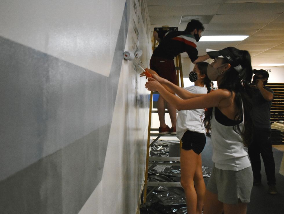 Two women with long brown hair and wearing masks, T-shirts and shorts use rollers to paint a wall with white paint. Behind then a young man in black shorts and a multi-colored shirt stands on a ladder.