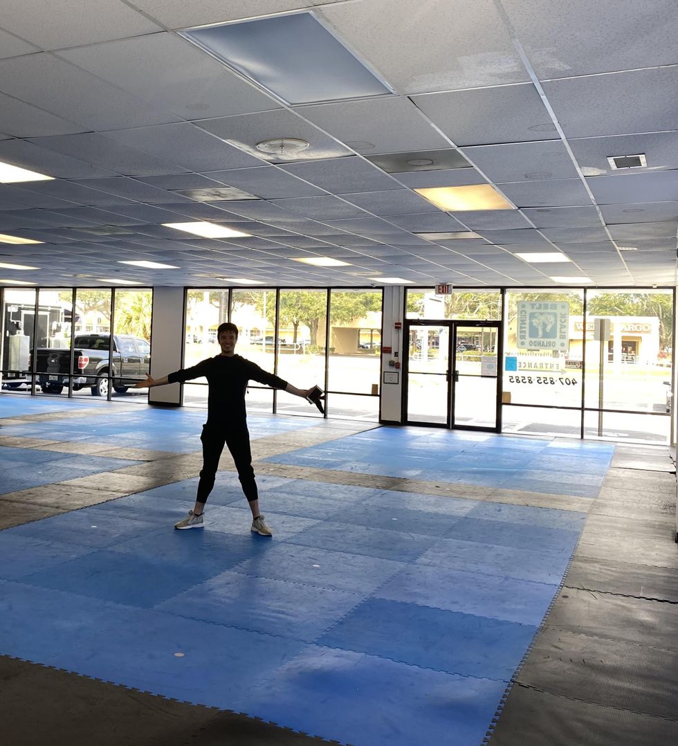 Inside a large, low-ceilinged but sunny studio space, Joseph Gatti spreads his arms and legs wide and smiles happily. The walls are covered in floor-to-ceiling windows and the black floor includes large squares of blue.