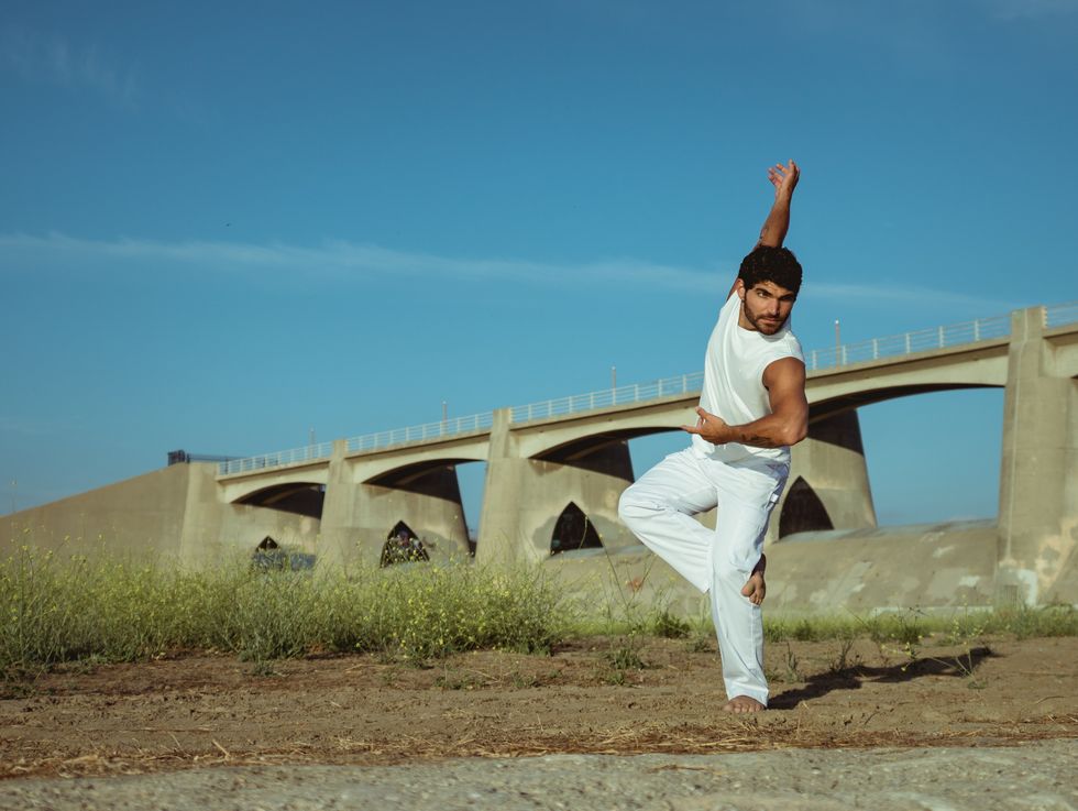 A male dancer in an all-white outfit dances barefoot outside in front of a bridge in Los Angeles.