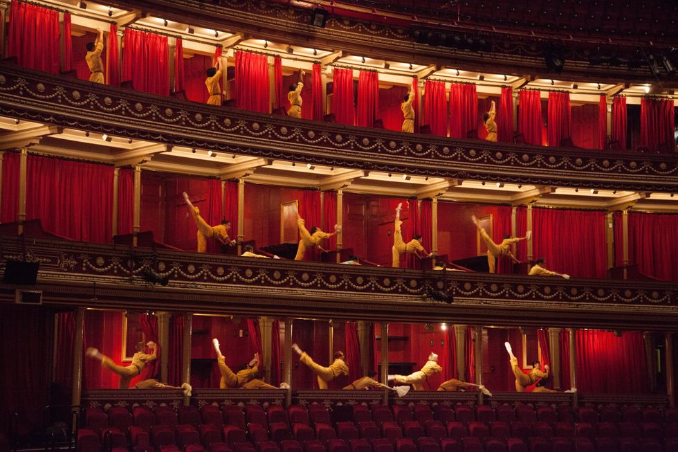 Dancers in tan jumpsuits penchu00e9 and reach upward throughout a rounded theater with red curtains and gold accents.