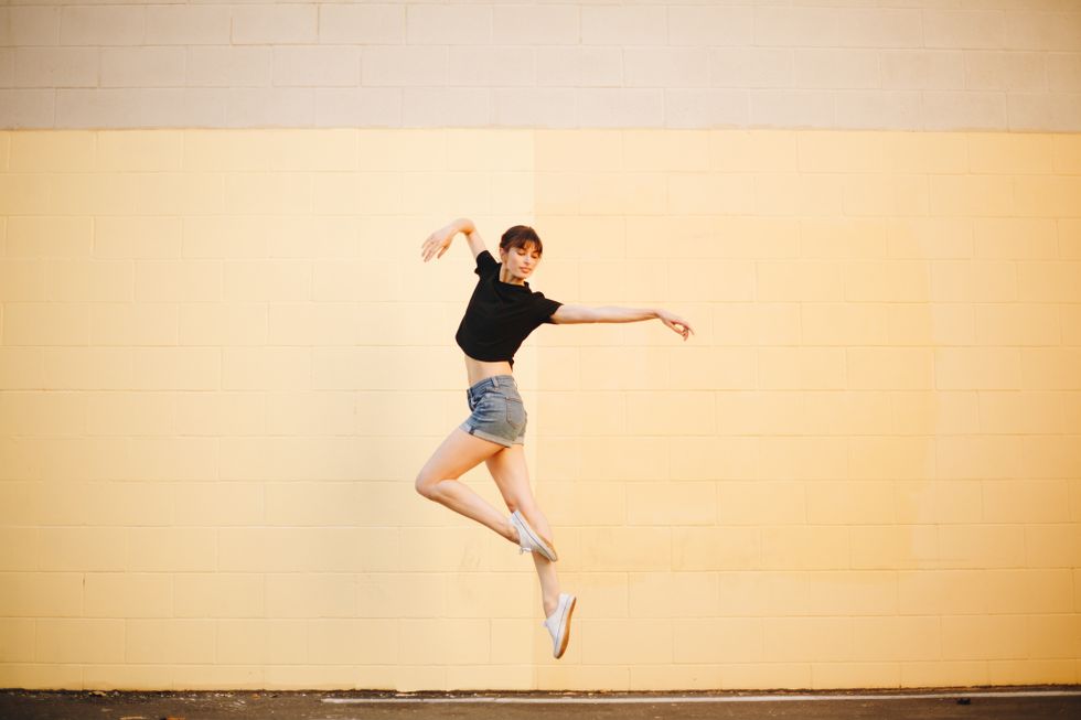 A female dancer in jean shorts and a black T-shirt skips outside in front of a yellow wall.