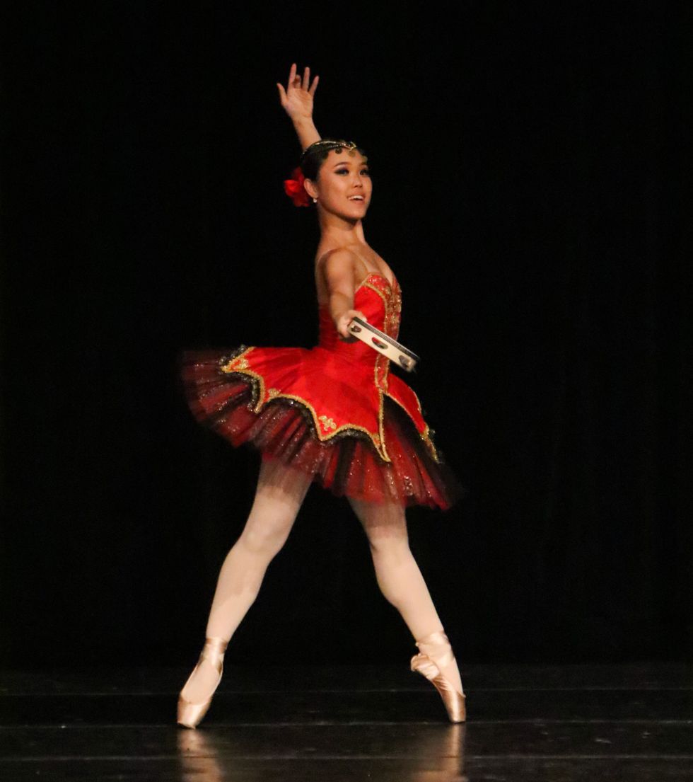 A young woman who seems to be performing the Kitri variation. She is in a slightly angled echappe on pointe, and wears a red and black tutu with a red hairpiece. She holds a tamborine which she extends out to the camera with one hand, the other arm behind her and up.