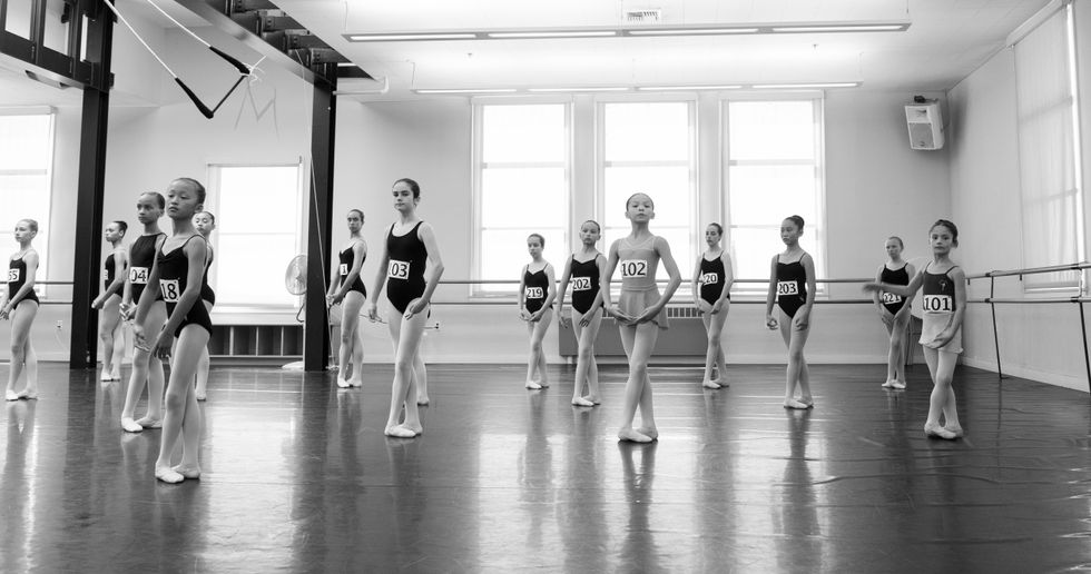 A master class of young girls. They each wear a number pinned to their leotard. They are in a large studio, standing in fifth position.